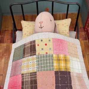 May include: A miniature bed with a black metal frame, featuring a patchwork quilt in various colours and patterns. A plush bunny head rests on the pillows, creating a cosy, inviting scene. Two yellow pillows with cross patterns complete the bed setting.