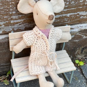 May include: A soft toy mouse with large ears, dressed in a beige knitted robe, seated on a miniature bench. The mouse wears a pink shirt under the robe. The bench has a light blue metal frame and a wooden seat.