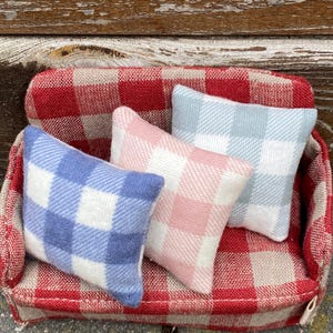 Może przedstawiać: Miniaturowa czerwono-biała kanapa w kratę z trzema poduszkami w kratę w kolorze niebieskim, różowym i jasnoniebieskim. Sofa i poduszki wykonane są z miękkiej, teksturowanej tkaniny. Poduszki mają kwadratowy wzór.