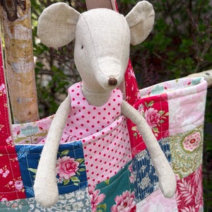 May include: A handmade fabric tote bag with a patchwork design in shades of red, pink, blue, and green, featuring floral patterns. A stuffed mouse doll with a pink polka-dot dress is inside the bag. The bag has a beige handle.