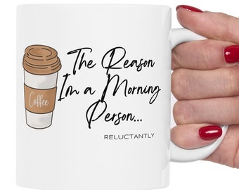 以下が含まれることがあります： 白いセラミック製のマグカップに、コーヒーカップの白黒のイラストと「The Reason I'm a Morning Person... Reluctantly.」というテキストが描かれています