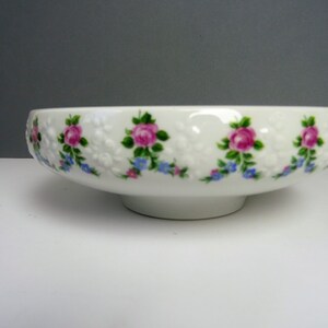 Könnte beinhalten: Eine weiße Keramikschale mit Sockel. Die Schale ist mit einem sich wiederholenden Muster aus rosa Rosen, grünen Blättern und blauen Blumen verziert. Der Rand hat ein strukturiertes, geprägtes Design.