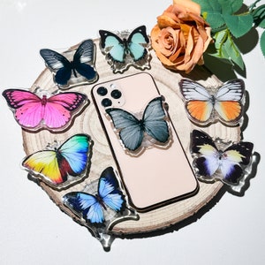 May include: A pink, yellow, blue, and black butterfly phone grip with a variety of butterfly designs on a wooden surface.