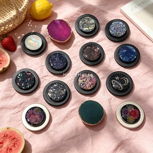 May include: A collection of circular phone grips featuring various designs including iridescent shells, dark glitter, floral patterns, and celestial motifs with stars and moons. The grips have black or silver bases and are arranged on a pink fabric surface.
