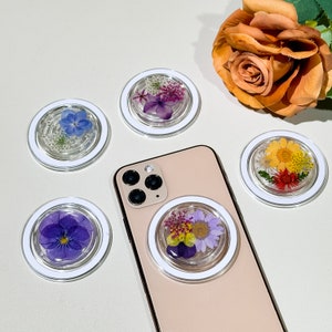 Puede incluir: Cinco agarres para teléfono con diferentes diseños de flores prensadas. Los agarres son blancos con un centro transparente y están unidos a una funda de teléfono dorada. La funda del teléfono está sobre una superficie blanca.