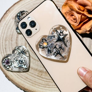 以下が含まれることがあります： 金色のiPhoneに、透明なハート型のスマホグリップが付いています。グリップには銀色のグリッターと、星と鍵の付いた銀色のチャームが付いています。