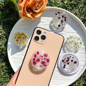 Könnte beinhalten: Eine weiße Handyhülle mit einem durchsichtigen Handygriff mit gepressten Blumen. Der Handygriff ist im Pop-Socket-Stil mit einem pink- und rotblumigen Design. Die Handyhülle liegt auf einer weißen Oberfläche mit anderen Handygriffen mit gepressten Blumen.