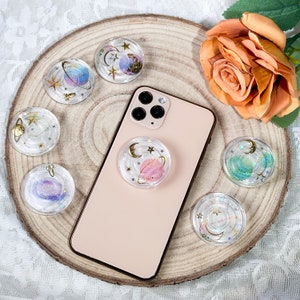 Puede incluir: Un agarre de teléfono rosa y dorado con un diseño de galaxia. El agarre está unido a una funda de teléfono rosa. Hay varios agarres de teléfono con temática de galaxia en la imagen, todos con diferentes colores y diseños.
