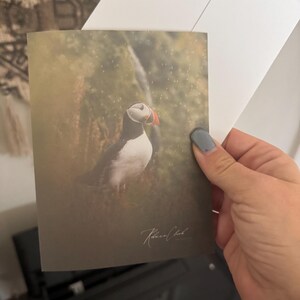 May include: A greeting card featuring a detailed illustration of a puffin bird with a white chest, black back, and a colorful beak. The card is held with a white envelope. The card has the signature "Karen Clark".