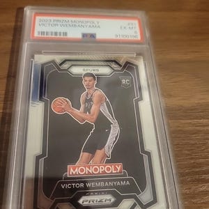以下が含まれることがあります： 2023年Prizm Monopoly Victor Wembanyamaバスケットボールカード、EX-MT 6評価、保護ホルダー入り。スパーズのユニフォームを着た選手がバスケットボールを持っているカード。MonopolyとPrizmのロゴが見えます。