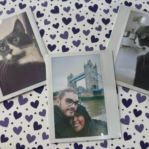 Puede incluir: Tres fotos instantáneas sobre una superficie blanca con un patrón de corazones morados. Dos fotos muestran gatos blancos y negros, y la tercera muestra a una pareja frente al Tower Bridge. Las fotos están en marcos blancos.