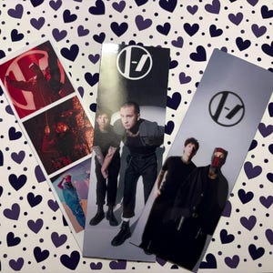 Può includere: Tre segnalibri rettangolari con immagini di un gruppo musicale. Ogni segnalibro ha un design diverso, che include i membri della band e il logo della band. Lo sfondo è un motivo di cuori viola su bianco.