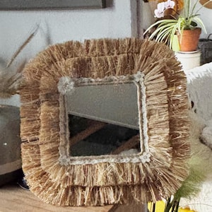 May include: Square mirror with a natural fiber frame. The frame has a layered, fringed design in a light brown color. The mirror is square with a white trim and small floral accents at each corner. Decorative home decor.