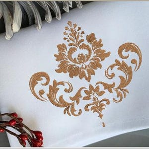 May include: White square decorative towel with a detailed gold damask pattern embroidered in the centre. The design features floral and scroll elements. The towel is folded, and the edges are visible. A red berry garland and decorative leaves are in the background.