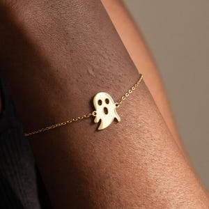 Op de afbeelding: Een goudkleurige armband met een centrale hanger in de vorm van een geest met uitgesneden ogen en een mond. De hanger is bevestigd aan een fijne, dunne metalen ketting die om de pols valt, met een gepolijste afwerking en een eenvoudig ontwerp.
