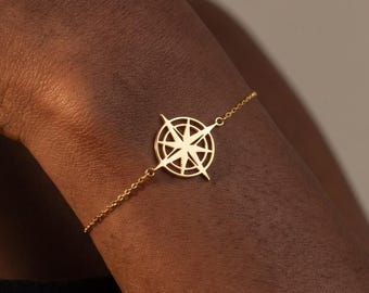 May include: A gold-toned bracelet featuring a central compass rose charm with a star-shaped design. The charm is connected to a delicate, thin chain that wraps around the wrist, showcasing a polished metallic finish and a symmetrical geometric shape.