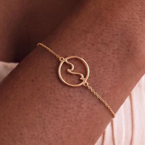 May include: A delicate gold chain bracelet featuring a circular pendant with an internal wave-like design. The bracelet is constructed from a thin, polished gold-tone metal chain that connects to both sides of the circular charm, creating a continuous loop.