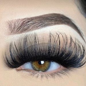 May include: Close-up of an eye with thick, black eyelashes and a well-defined, dark brown eyebrow. The eye has a light blue iris with a brown centre. The skin around the eye is a light beige colour.