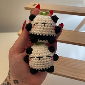以下が含まれることがあります： 手作りのパンダのキーホルダー2つ。各パンダは、黒いアクセント、黒い足、中央に黒いストライプが付いた白い体を持っています。彼らは目を閉じ、ピンクの頬、そして上に小さな緑の葉を持っています。