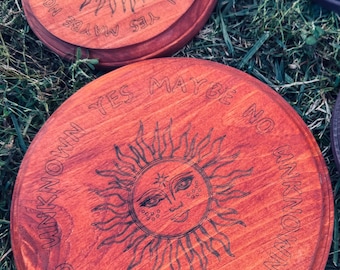 May include: Wooden coasters with a sun design and the words "Yes Maybe No Unknown" etched around the edge. The coasters are reddish-brown and sit on green grass.