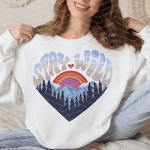 Puede incluir: Sudadera blanca con un gráfico en forma de corazón que dice "Stay Wild" en una fuente retro. El gráfico presenta un arcoíris, montañas y pinos.