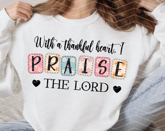 Peut inclure: Sweat-shirt blanc avec le texte "With a thankful heart, I PRAISE THE LORD" en lettres noires. Le mot "PRAISE" est dans des carrés colorés avec des motifs floraux.