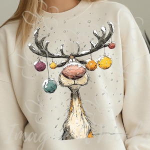 Puede incluir: Sudadera color crema con un dise&ntilde;o caprichoso de reno de dibujos animados. El reno tiene grandes astas adornadas con adornos coloridos y una prominente nariz roja. Los copos de nieve est&aacute;n esparcidos alrededor del reno, creando una escena invernal festiva.