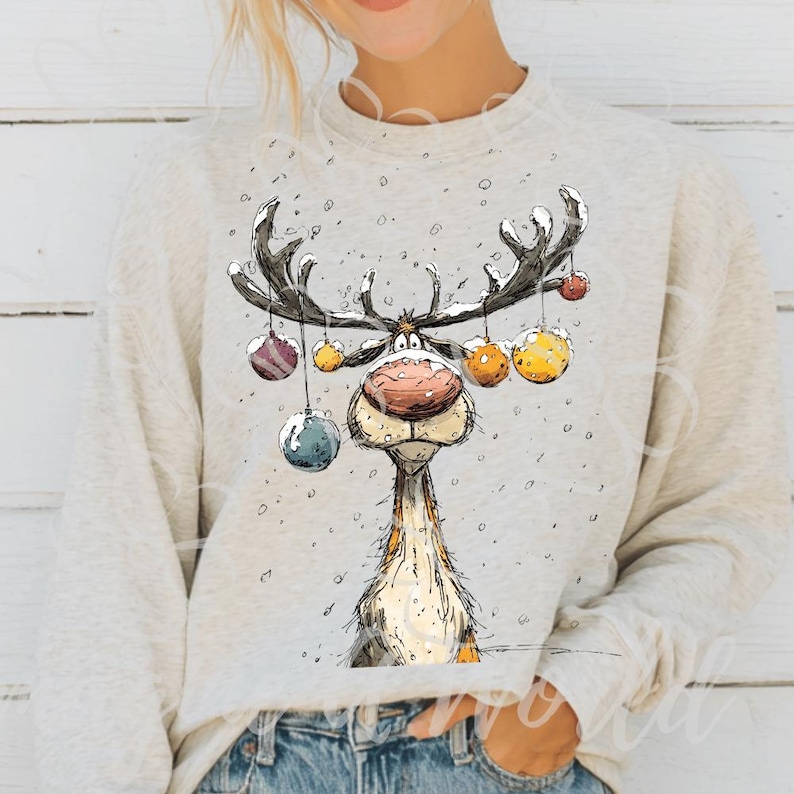 Puede incluir: Sudadera gris claro con un dise&ntilde;o de reno de dibujos animados. El reno tiene grandes cuernos adornados con bolas de colores y una nariz roja. El dise&ntilde;o est&aacute; sobre un fondo nevado, perfecto para la temporada navide&ntilde;a.