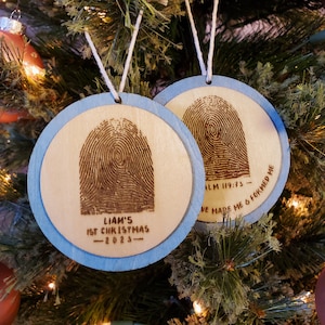 May include: Two round wooden ornaments with blue trim. One ornament has a fingerprint design and the text "LIAM'S 1ST CHRISTMAS - 2023". The other ornament has a fingerprint design and the text "PSALM 119:73 - YOU HAVE MADE ME & FORMED ME".