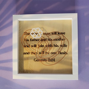 May include: A framed print with a gold background and a white geometric design. The print features the text "The man will leave his father and his mother and will join with his wife and they will be one flesh. Genesis 2:24".