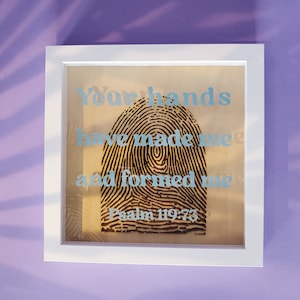 May include: Framed artwork featuring a large fingerprint design in black and brown. The text "Your hands have made me and formed me" is in light blue, along with "Psalm 119:73". The white frame contrasts with the artwork.