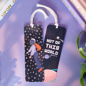 May include: Two black bookmarks with white string ties. One bookmark features a woman in a spacesuit floating in space with a starry background. The other bookmark has the text "Not of this world" with a bible verse and a planet illustration.