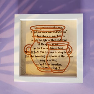May include: A framed print with a beige background and a copper-colored design of two vases with a Bible verse from 2 Corinthians 4:6-7. The verse reads: "For God, who said, "Let light shine out of darkness," has shone in our hearts to give the light of the knowledge of the glory of God in the face of Jesus Christ. But we have this treasure in jars of clay to show that this all-surpassing power is from God and not from us."