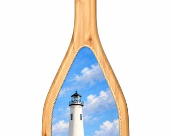 May include: A decorative wooden paddle featuring a light brown wood grain finish with a rope-wrapped handle. The blade displays a printed scene of a white lighthouse on a sandy beach with blue sky, clouds, and ocean waves.