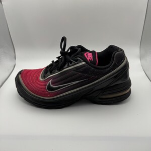 May include: Black and red athletic shoe with the Nike logo in pink. The shoe features a black sole, a red toe, and a black upper with a silver swoosh. The shoe has black laces.