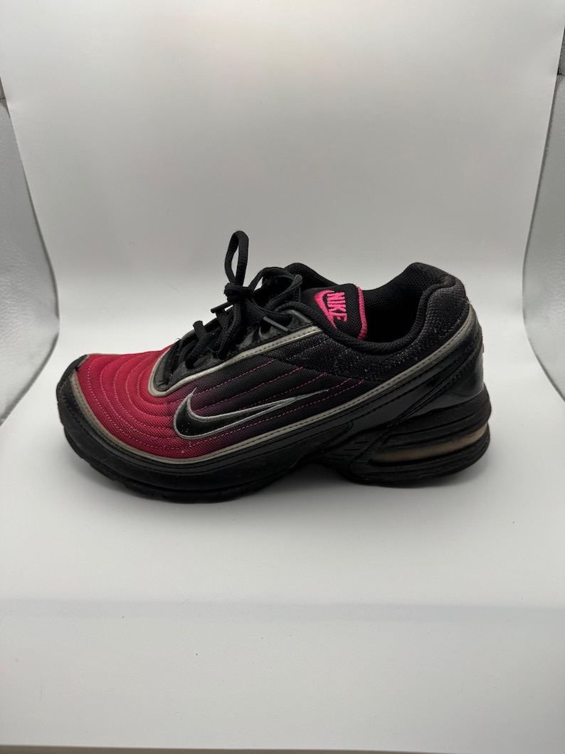 May include: Black and red athletic shoe with the Nike logo in pink. The shoe features a black sole, a red toe, and a black upper with a silver swoosh. The shoe has black laces.