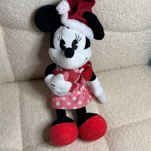 Op de afbeelding: Een Minnie Mouse pluche speelgoed met een rode kerstmuts met witte rand. Het speelgoed heeft zwarte oren, een wit gezicht en een roze polkadot jurk. Rode strik en schoenen maken de feestelijke look compleet.