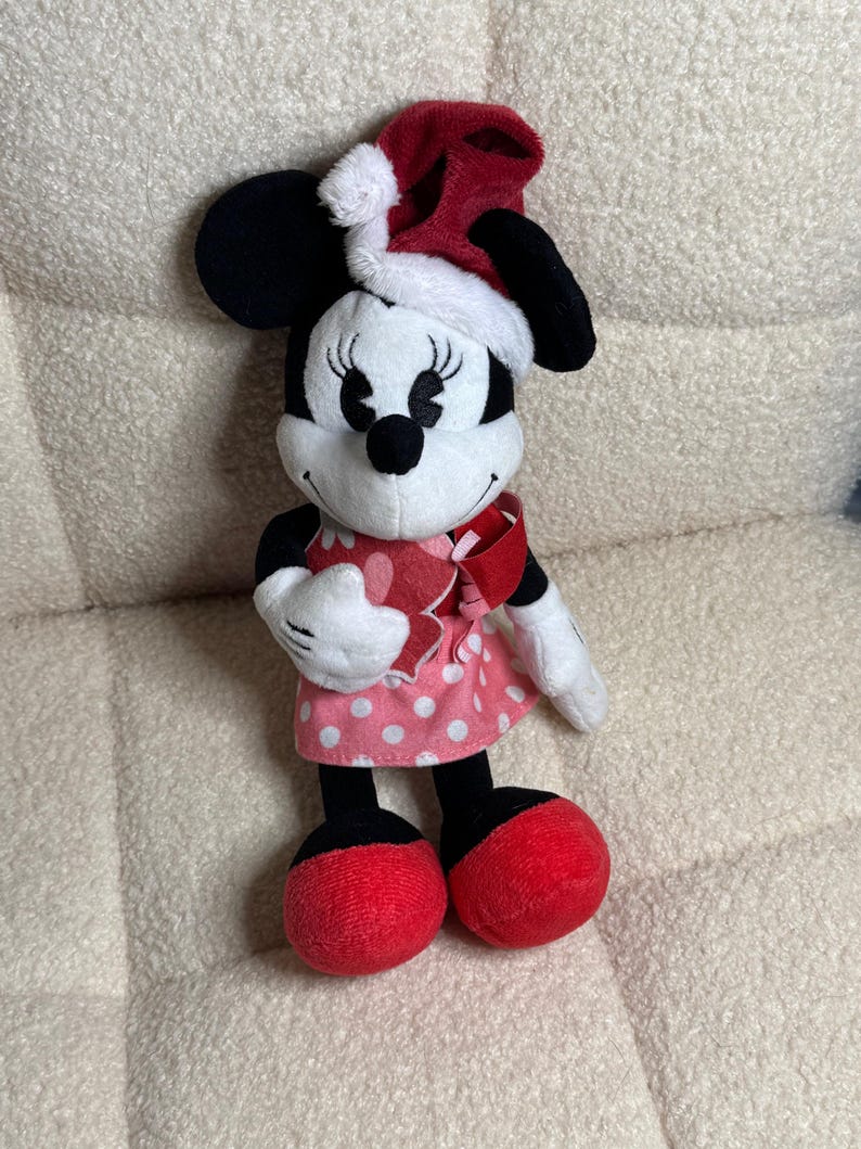 Op de afbeelding: Een Minnie Mouse pluche speelgoed met een rode kerstmuts met witte rand. Het speelgoed heeft zwarte oren, een wit gezicht en een roze polkadot jurk. Rode strik en schoenen maken de feestelijke look compleet.
