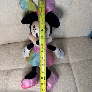 May include: A Minnie Mouse plush toy, approximately 15 inches tall, with a rainbow-colored dress and bow. The toy has black ears, a white face, and pink shoes. A yellow measuring tape is held next to the toy.