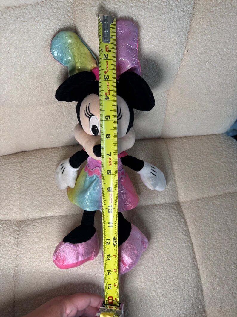 May include: A Minnie Mouse plush toy, approximately 15 inches tall, with a rainbow-colored dress and bow. The toy has black ears, a white face, and pink shoes. A yellow measuring tape is held next to the toy.