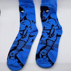 May include: A pair of blue socks with a black graphic design. The design appears to be a stylized superhero figure. The socks are made of a soft material and are likely for everyday wear.