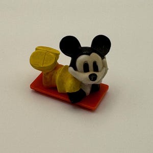 May include: A small Mickey Mouse figurine, lying on a red rectangular base. The figure has black ears, a white face, and yellow shorts. The figurine is a collectible item.