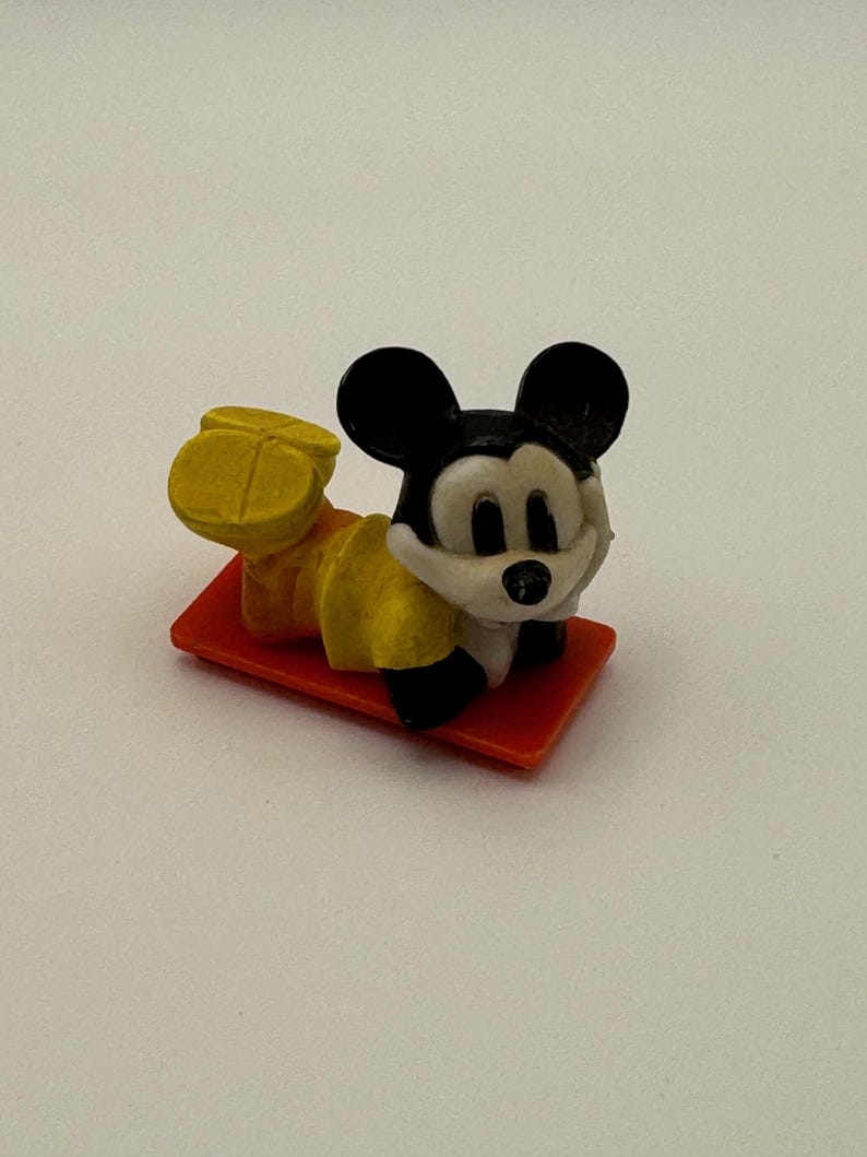 May include: A small Mickey Mouse figurine, lying on a red rectangular base. The figure has black ears, a white face, and yellow shorts. The figurine is a collectible item.