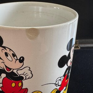 May include: White ceramic mug with a cartoon image of Mickey Mouse and a smaller Mickey Mouse on the side. The mug has a chip on the rim.