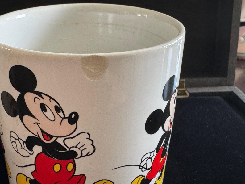 May include: White ceramic mug with a cartoon image of Mickey Mouse and a smaller Mickey Mouse on the side. The mug has a chip on the rim.