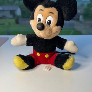 May include: A vintage Mickey Mouse soft toy. The toy has a black body, ears, and tail, with a tan face and white hands. It wears red shorts with yellow buttons and yellow feet. The toy is sitting on a white surface.
