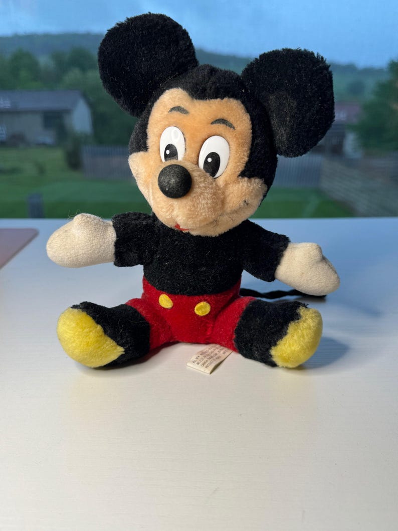 May include: A vintage Mickey Mouse soft toy. The toy has a black body, ears, and tail, with a tan face and white hands. It wears red shorts with yellow buttons and yellow feet. The toy is sitting on a white surface.