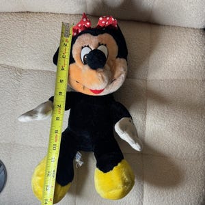 Puede incluir: Mu&ntilde;eca de peluche de Minnie Mouse, de aproximadamente 38 cm de altura, con cuerpo negro, pies amarillos y guantes blancos. Tiene un lazo rojo con lunares blancos y una cara sonriente con boca roja. La mu&ntilde;eca es de tela suave.