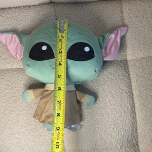May include: A plush toy of a character with large black eyes, green skin, and large ears with pink insides. The toy is approximately 11 inches tall. It is wearing a tan outfit and has small hands and feet.