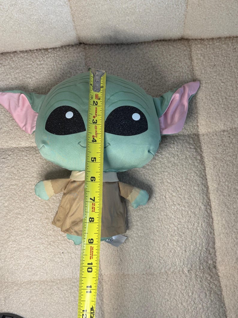 May include: A plush toy of a character with large black eyes, green skin, and large ears with pink insides. The toy is approximately 11 inches tall. It is wearing a tan outfit and has small hands and feet.